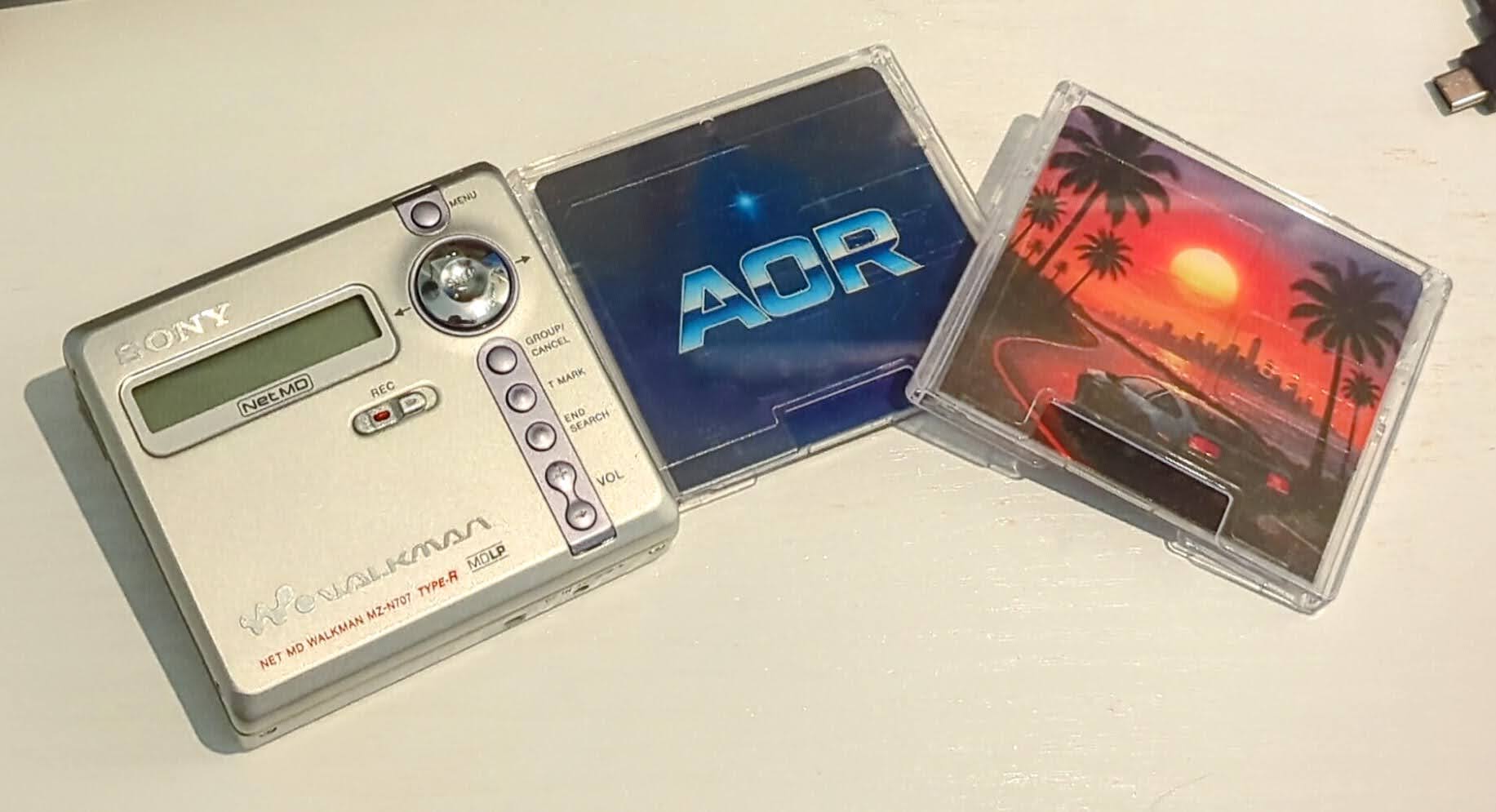 A Minidisc player and two UV-printed discs, one with a car driving in the sunset and another with the letters AOR.